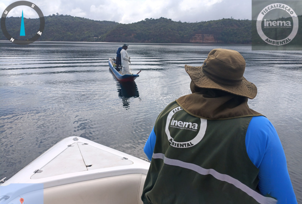 Fiscal ambiental do INEMA em um barco observando dois pescadores em outra embarcação durante uma operação de fiscalização em um lago cercado por vegetação e montanhas.