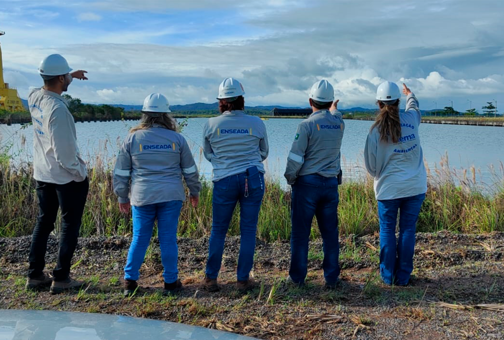 Grupo de profissionais da área ambiental, vestindo uniformes com logotipo "Enseada" e capacetes, observando e analisando uma área de recurso hídrico. O cenário inclui vegetação ao redor do corpo d'água e céu parcialmente nublado.